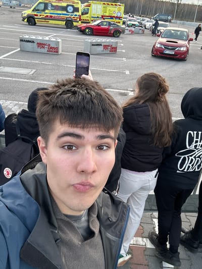 The image depicts an outdoor automotive event in Wolica, Poland, captured from a first-person perspective in a selfie format. In the immediate foreground, a young male with short brown hair and brown eyes is taking a selfie, looking directly at the camera with slightly pursed lips. He is wearing a dark blue outer jacket over a brown sweater.

Behind the selfie-taker, another individual's hand is visible holding a black smartphone vertically, capturing a scene beyond. To the right of the selfie-taker, partially obscured, is a female with long brown hair, wearing a black puffer jacket and light-colored pants. Further to the right, another person is partially visible, wearing a black hoodie with white text on the back, partially legible as "CHA... OBR...".

The background features a large, paved asphalt area, likely a parking lot or track, with white painted markings. Parked centrally are two yellow ambulances; the left one bears "AMBULANS" and a red cross-like emblem, while the right one shows "PAŃSTWOWE RATOWNICTWO MEDYCZNE" and a Star of Life symbol. Between and in front of the ambulances, a red sports car (resembling a Nissan S-chassis) with significant front-end damage, widebody fender flares, and green/yellow accented wheels is parked.

Further into the background, a red Subaru Impreza, possibly a WRX or STI model, is visible with a Polish license plate "WP 1078Z" on its lower front bumper. Multiple other vehicles are queued or parked in the distance. Several large, grey concrete barriers, resembling LEGO blocks, are positioned on the asphalt. These barriers display the text "MANNOL" and "FUNDACJA FUN-DRIVE DZIECIOM" (Fun-Drive Foundation for Children). The sky is overcast, and sparse trees are visible in the far background.