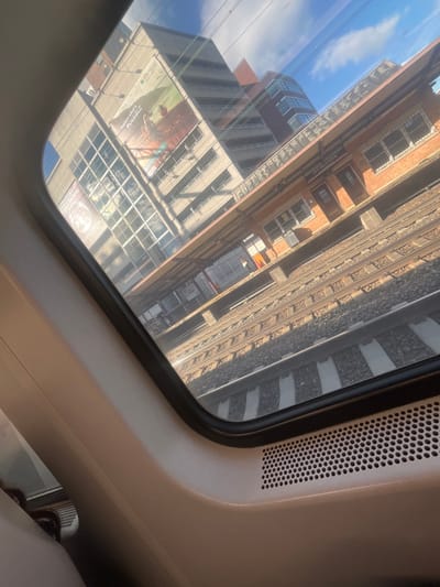 A view through a train window captures a cityscape. Buildings with numerous windows reflect the sky. Adjacent to the buildings, train tracks stretch into the distance. The window frame is visible, with ventilation holes near the top right corner. The interior of the train is partially visible in the bottom right corner. The scene presents an unusual perspective of urban life alongside rail transportation.