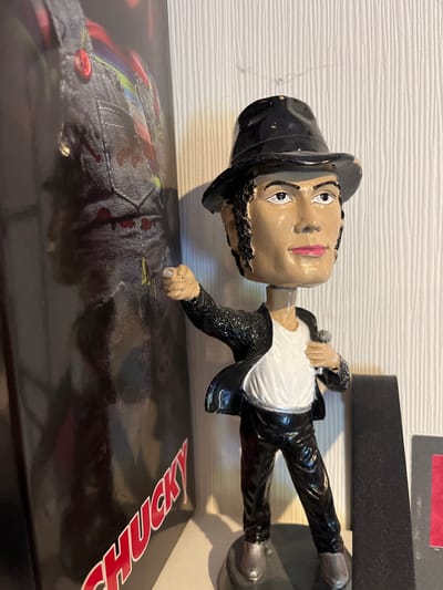 A Michael Jackson bobblehead is positioned to the right of a tall, dark object with the word "CHUCKY" in red lettering, which appears to be a box or poster. The bobblehead is wearing a black fedora, a black sparkly jacket over a white t-shirt, and black pants. It is holding a microphone in its left hand and pointing with its right index finger. The background consists of a textured, off-white wall and a dark vertical object to the right of the bobblehead. The scene is indoors, likely on a shelf or display. The location is not specified to be in Schwäbisch Hall, Germany.