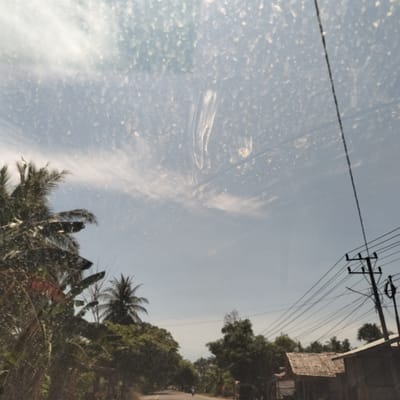 The image shows a view from inside a vehicle, likely a car, looking out at a rural landscape in Aluekeujruen, Indonesia. The windshield is dirty, with water spots and streaks obscuring the view.

Outside, the scene depicts a road lined with lush tropical vegetation, including palm trees. An unidentifiable person is riding a motorcycle on the road. A utility pole with numerous power lines is visible on the right side of the frame, along with a small hut-like structure with a thatched roof. The sky is light blue with some wispy clouds.