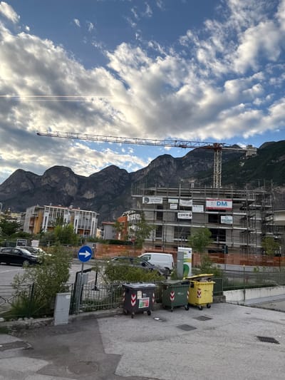 This image depicts a construction site with a large crane prominently featured against a backdrop of a mountain range and a cloudy sky. The scene is oriented vertically. The construction site appears to be in an urban or semi-urban setting, with several buildings under construction, some partially clad in scaffolding and orange safety netting. Signs are visible on the scaffolding, including "DM," "MA CO.S.," "GIRARDI," "ESSEBI," and "GRUPPO DE VIALI."

In the foreground, to the right, are several large waste bins. One is yellow, and two are dark green and grey, respectively, with reflective arrows on their sides. A white van and other vehicles are parked along the road. A blue traffic sign with a white upward-pointing arrow is visible. The overall impression is of an active construction project in a mountainous region.

The time of day appears to be late afternoon or early evening, given the angle of the sun and the quality of light illuminating the clouds. The sky is a mix of blue sky and substantial white and grey clouds, suggesting varied weather. The location is identified as Mezzolombardo, Italy, which is consistent with the mountainous scenery and the presence of Italian text on the signs. No people are clearly visible in the image.
