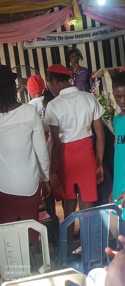 The image captures an indoor event, likely a religious service or gathering, taking place in Agbara, Nigeria. Several individuals, primarily young people, are visible. In the foreground, two individuals are partially visible, one wearing a white long-sleeved shirt and a red skirt, and another in a white short-sleeved shirt and a red skirt. Behind them, another person in a white shirt and red skirt is also present. To the right, a young male is wearing a teal shirt with text that reads "EM". At the back, a woman in a purple dress is speaking into a microphone, possibly addressing the gathering. A banner with text that reads "Jesus Christ The Same Yesterday, And Today, And Fo" and "HEB. 13:8" is displayed prominently on the wall. The environment appears to be decorated with draped fabric in pink and yellow hues, and a striped black and white background is visible. Several plastic chairs are scattered throughout the scene, some of which have writing on them. The overall atmosphere suggests a formal or semi-formal event. The timestamp on the image indicates it was taken on June 28, 2026, at 09:29.