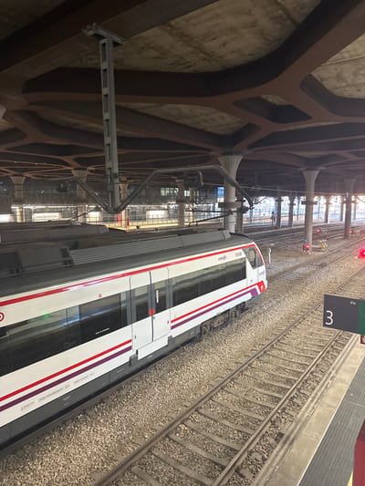 The image shows a Renfe train, number 515M, at a station in Oviedo, Spain. The train is white with red and purple stripes. It is parked on a track next to a platform. A sign indicates "Vía 3". The station has a large, modern roof structure supported by pillars. There are other tracks visible in the background, with overhead electrical lines. The ground is covered with gravel and ballast. The lighting suggests it is daytime, though the station is covered. No people are clearly visible.