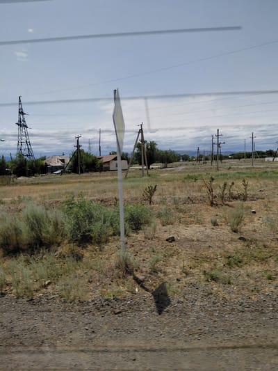 The image depicts a rural or semi-rural landscape with power lines and utility poles dominating the sky. In the background, several houses and what appear to be farm buildings are visible, clustered together amidst scattered trees. A large, industrial-looking tower is prominent on the left side of the frame. The foreground is characterized by dry, sparse vegetation and a dirt or gravel road. The sky is partly cloudy with a muted, overcast appearance. The overall scene suggests a utilitarian and somewhat undeveloped area, possibly on the outskirts of a settlement like Ақтүбек, Kazakhstan, given the context provided. There are no people visible in the image.
