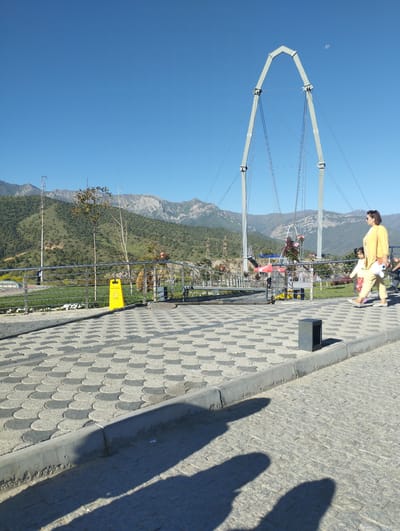 In Dardar, Uzbekistan, a paved walkway with a decorative pattern of gray and off-white scales leads towards a park area. A woman in a yellow outfit and a young child in a similar color, also with a yellow top and patterned leggings, walk along the walkway. Behind them, a metallic arch structure, possibly part of a bridge or attraction, is visible against a clear blue sky with a crescent moon. The park features a metal railing, green grass, trees, and a backdrop of rolling, green, and rocky mountains under a bright sun. A yellow hazard sign is placed near the railing. Shadows of people and possibly a structure are cast on the patterned walkway.