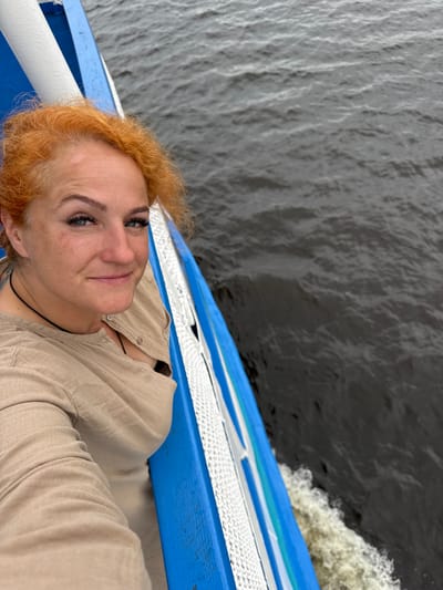 A woman with red, curly hair is taking a selfie on a boat. She is wearing a tan, button-up shirt and a black necklace. She has blue eyes, makeup, and a light tan complexion. The boat has a blue and white railing. The water is dark gray and choppy, indicating the boat is moving. The scene is likely outdoors, on a body of water.