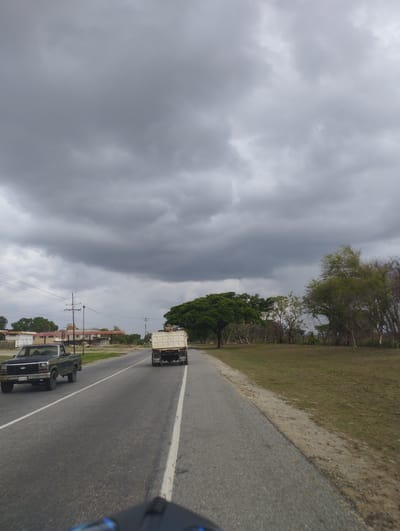 The image captures a road scene on an overcast day in El Bajío, Venezuela, likely from the perspective of a moving vehicle, indicated by the top of a helmet or dashboard in the bottom foreground. The sky is dominated by thick, dark grey clouds, suggesting impending rain or heavy cloud cover.
A two-lane asphalt road stretches into the distance, featuring a broken white center line and a solid white line marking the right shoulder. On the left side of the road, a dark-colored (possibly dark green or black) pickup truck with a chrome bumper is visible, moving in the same direction as the viewer. Further ahead on the right lane, a large, light-colored dump truck is seen from the rear, also traveling forward. Visible on the mudflaps of the dump truck are the words "BATT MARK" and "MADE IN USA".
The landscape alongside the road is rural to semi-rural. On the left, beyond the road, there are some buildings, possibly part of a small town or settlement, along with utility poles and power lines. Further down the road on the left, dense green trees line the area. The right side of the road features a grassy field and a mix of trees, some appearing lush green and others more sparse or dormant. The overall atmosphere is subdued due to the heavy cloud cover.