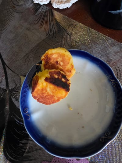 Two golden-brown, fried patties, likely syrniki or pancakes, rest on a white plate with a blue rim. The patties show signs of being slightly overcooked in some areas, with darker brown char marks. They are positioned in the center of the plate, which is itself placed on a patterned tablecloth with dark grey and gold accents. The background is slightly blurred, showing a wooden surface and hints of white crocheted material. The scene appears to be an indoor setting, likely a dining table. There is no indication of the city of Nevzorovo, Russia, within the image itself.