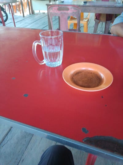 A close-up shot on a red table surface in Glee Gogo, Indonesia, captures a clear glass mug filled with a pale liquid and an oval-shaped orange plate containing a brown powder, possibly coffee or spice. In the background, a blurry glimpse of outdoor seating with orange and red chairs and a wooden floor suggests a casual dining or rest area. A person's dark-clothed legs are visible at the bottom of the frame, indicating their presence at the table. The lighting appears natural, casting soft shadows.