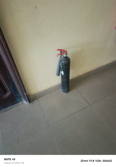 The scene, captured indoors in Gwammaja, Nigeria, on 2026.01.27 at 02:02 PM, depicts a black, cylindrical fire extinguisher standing upright against a pale yellow wall. The extinguisher features a red handle assembly with a visible red safety pin and a black discharge nozzle. White scuff marks are present on the extinguisher's body. A dark baseboard runs along the bottom of the wall, where it meets a tiled floor. The floor consists of dark gray square tiles with visible grout lines. On the left side of the frame, a dark brown door frame is partially visible, exhibiting wear and light discoloration at its base. No individuals, actions, or interactions are observed within the frame.