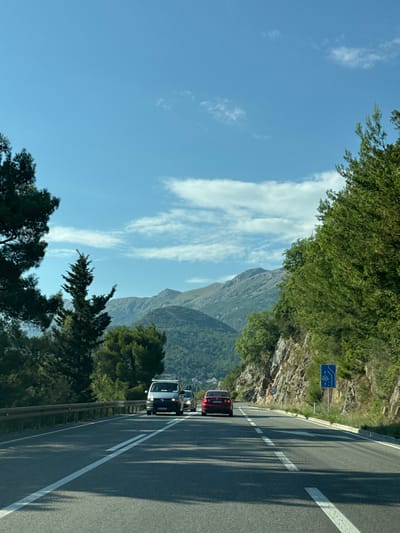 The image shows a road with cars driving through a mountainous landscape. In the foreground, the asphalt road has white lane markings. A gray van is driving in the left lane, followed by a red car. On the right side of the road, a rocky cliff face is visible, covered in vegetation. A blue traffic sign with white symbols indicating a lane closure is mounted on a pole next to the road, along with a speed limit sign showing "50". In the background, rolling green mountains are visible under a clear blue sky with scattered white clouds. The overall scene suggests a scenic drive on a sunny day. The location is identified as Košljun, Montenegro.