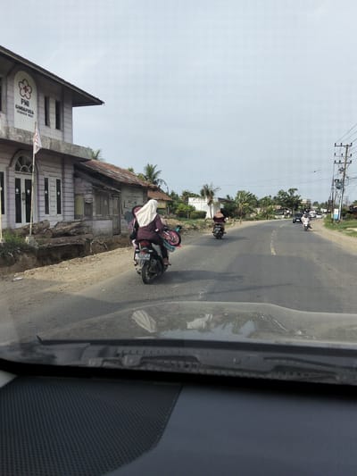 A street scene in Keudelapang, Indonesia, shows a road with several motorcycles and some cars. In the foreground, the interior of a vehicle is visible, including the dashboard and windshield. On the left side of the road, a building with a sign that reads "PMI GANDAPURA BIREUEN NAD" is present. A motorcycle with two individuals, one driving and one passenger, is moving away from the camera on the left side of the road. Another motorcycle is further down the road on the right. The sky is overcast.