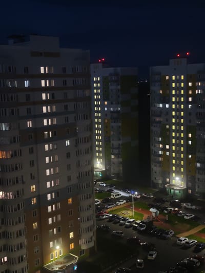 The image shows an aerial view of a residential complex at night, likely in Kursk, Russia. Three tall apartment buildings are visible, with many windows illuminated, suggesting residents are present. Red lights are flashing on top of two of the buildings, possibly for aviation safety. Below the buildings, a parking lot is filled with numerous cars, and a playground area is also visible. The overall scene depicts a quiet, urban residential setting after dark.