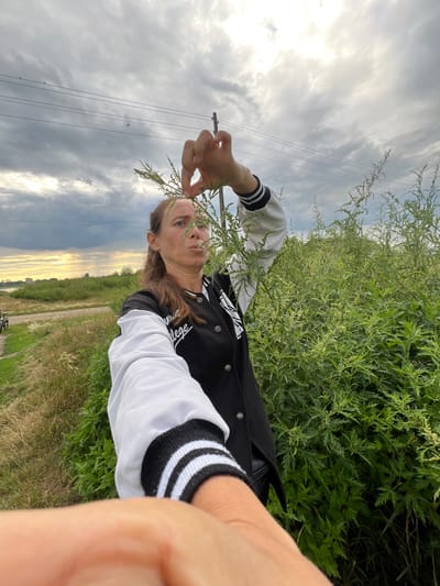 A woman stands in a field of tall green grass and weeds, wearing a black and white letterman jacket. She holds a sprig of greenery up to her face, her expression neutral. The sky above is cloudy with patches of sunlight breaking through, suggesting it might be late afternoon or early evening. A rural landscape with a dirt road and power lines can be seen in the background. The image is oriented vertically. No visible text.