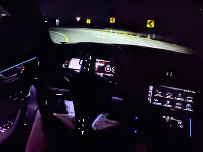 This image captures the interior of a car at night, likely in Bezden, Bulgaria, as suggested by the user prompt. The driver's perspective is shown, with the steering wheel and dashboard illuminated by ambient light and the car's internal lighting. The road ahead is visible through the windshield, curving to the right and marked with yellow lines. Several road signs are visible, including a yield sign, two chevron signs indicating curves, and a digital heads-up display projected onto the windshield showing navigational information and speed.

The dashboard features multiple screens, including a central infotainment display showing various icons for functions like radio, media, navigation, and phone. Below this, another screen displays climate control information, showing a temperature of 70 degrees. Blue LED ambient lighting is present along the dashboard and door panels. A person's legs are visible in the lower left of the frame, indicating the presence of a driver or passenger. The overall environment is dark, consistent with nighttime driving.