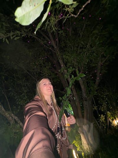 A low-angle medium shot shows a young Caucasian woman from the chest up, looking up and to her left with a slight smile. She has blonde hair and is wearing a dark brown satin shirt and a pearl necklace. In her right hand, she is holding a dark cylindrical object with an orange label. The background is a dark, wooded area with trees and foliage. Small, purple lights are scattered throughout the branches of the trees, suggesting some form of decoration or natural phenomenon. The lighting is dim, indicating it is nighttime. The location is Zavod Mikhailovskii, Russia.