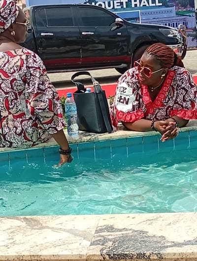 Two women are seated outdoors beside a turquoise water feature, likely a fountain or a shallow pool, in what appears to be Unguwan Sanusi, Nigeria. The scene is captured during the daytime under bright lighting.

On the left, a woman seen from behind is wearing a patterned red, white, and brown traditional garment (likely an Ankara print) and a matching headwrap. She has her right foot, adorned with dark ankle beads, partially submerged in the water, causing ripples. A black handbag and two plastic water bottles are visible on the edge of the water feature next to her.

On the right, another woman is partially visible from the chest up, leaning forward with her hands clasped. She has reddish-brown braided hair pulled back, and is wearing oversized red-rimmed sunglasses and a patterned traditional top with red trim and white accents, similar in style to the first woman's attire. A gold necklace and bracelet are also visible. On her top, there's a white patch with partial text that appears to read "202...", "Kaduna S...", and "EN", possibly indicating an event or organization related to Kaduna State.

In the background, a dark-colored pickup truck (resembling a Toyota Hilux) is parked. Behind the truck, a large white building is visible with the words "BOUQUET HALL" written on its facade, suggesting the setting is an event venue. The water feature has a light-colored, possibly granite or marble, tiled edge.