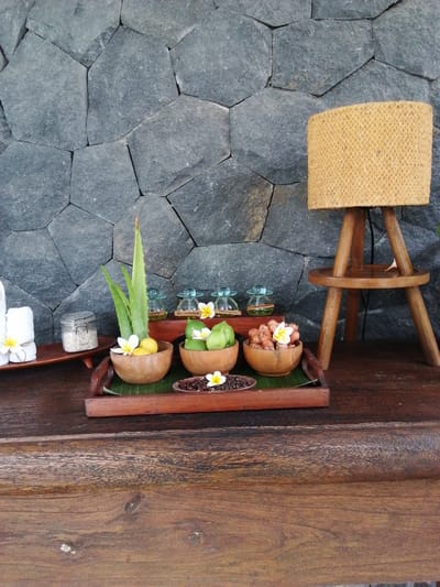 A spa-like arrangement is set against a stone wall. On the left, two white towels are folded and tied with a yellow flower. Next to the towels is a small glass jar with a metal lid, filled with white granular material. In the center, a wooden tray holds several small wooden bowls containing natural spa ingredients. One bowl contains a sprig of aloe vera, a yellow fruit, and a white flower. Another bowl has green fruits, and a third holds dried roots and a white flower. A small dish with dark peppercorns and a white flower sits in front of the bowls. Behind the tray, a series of small glass bottles with green liquid are lined up. To the right, a woven lampshade is mounted on a wooden tripod. The entire arrangement rests on a dark wooden surface. The scene is likely from a spa or resort in Banyuwangi, Indonesia, given the natural elements and tropical ambiance.