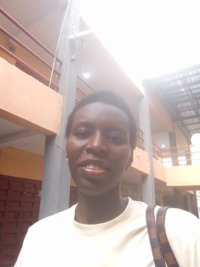 A brightly lit, outdoor portrait captures a young Black person, likely female, from the chest up, smiling directly at the camera. She has short, dark, natural hair and a wide, cheerful smile revealing white teeth. She is wearing a plain, light-colored (possibly off-white or cream) short-sleeved t-shirt and small, silver-colored stud earrings. A brown and dark brown checkered strap, likely from a shoulder bag, is visible over her right shoulder.
The setting appears to be a multi-story building, likely outdoors or in an open courtyard, consistent with an urban environment like Satellite Town, Nigeria. The background features structural elements such as light orange-peach colored walls and beams, grey support pillars, and dark reddish-brown metal railings. On one of the grey pillars, a small white satellite dish is mounted, along with some visible wiring. Other parts of the building, including a dark, possibly metal, roof or covering, are visible in the upper right. The strong, direct lighting suggests it is daytime, with some areas of the background appearing overexposed due to the brightness. The person's expression conveys happiness or contentment. No discernible text is visible in the image.