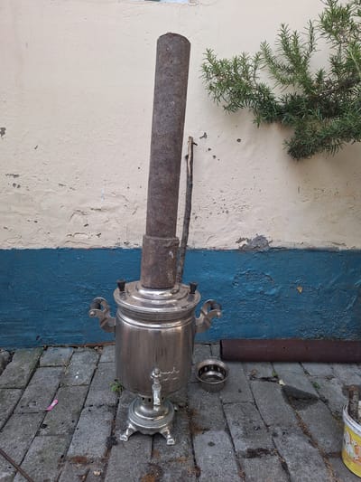 In Nabran, Azerbaijan, a weathered, silver samovar stands on a cobblestone patio, its tall, rusted chimney pipe extending upwards. The samovar has ornate handles and a spigot. A small, metal strainer sits on the ground next to it. Behind the samovar, a wall is painted in two horizontal bands: the upper section is a faded cream, textured and peeling in places, while the lower section is a vibrant, textured blue. A sprig of green rosemary grows from the cream section of the wall, its branches reaching towards the right. To the right of the samovar, a rusty metal pipe lies horizontally on the ground. In the lower right corner, a yellow bucket with a floral pattern contains a bundle of sticks.