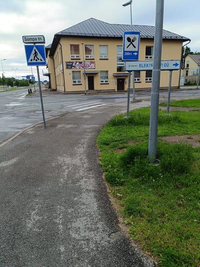 In the city of Jõhvi, Estonia, an outdoor scene captures a street corner with a yellow building. A paved sidewalk curves towards the right, bordered by a strip of green grass. Across the street, a two-story yellow building with a dark roof features multiple windows and doorways. Several street signs are visible: one indicates "Sompa tn." and a pedestrian crossing, another displays a fork and knife symbol with "200m" and an arrow pointing to the right, and a third sign reads "elektriteenindus ELEKTERVII OÜ" with an arrow pointing right. A pedestrian crossing is marked on the asphalt. The sky is overcast, and streetlights are present along the road. The overall environment appears to be a commercial or mixed-use area.