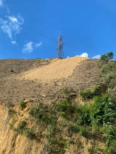 The image depicts a steep, barren hillside with a large pylon structure and power lines against a blue sky with scattered white clouds. The hillside appears to have been recently cleared of vegetation, with patches of loose, light-colored soil and small stones dominating the upper slope. Stumps and dead branches from former trees are visible scattered across the lower, darker brown sections of the slope. Some sparse green vegetation, including grasses and small bushes, is growing along the bottom and right side of the hillside, indicating some regrowth. The power lines stretch across the top of the frame, connecting to the pylon which is positioned on the upper part of the slope. No people or animals are present in the scene. The overall impression is one of land disturbance and the presence of electrical infrastructure. The scene is located in Thôn 18, Vietnam.