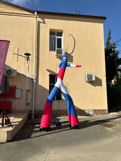 A tall, inflatable figure in Russian flag colors, red, white, and blue, stands in front of a pale yellow, two-story building. The figure is positioned on asphalt, with its long legs spread wide. The building has several windows, some with white frames and dark panes, and two air conditioning units mounted on the exterior wall. A banner with pink lettering, partially obscured by shadow, is visible on the left side of the image. Electrical wires and an antenna are mounted on the building. The scene appears to be outdoors, with a clear blue sky and shadows cast by the building and the inflatable figure, suggesting it is daytime. The overall impression is of an outdoor event or advertisement. The presence of the Russian flag colors on the inflatable suggests a connection to Russia, and the "Прогресс" signage on the banner points to the city of Progress, Russia.