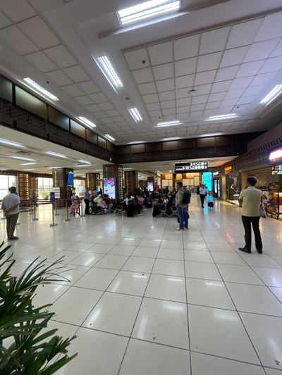 The image depicts the interior of an airport terminal, likely in Ahmedabad, India, given the context. The scene shows a spacious, well-lit hall with a high, tiled ceiling featuring numerous fluorescent light fixtures. The floor is tiled with large, light-colored squares, reflecting the bright overhead lighting.
In the foreground, a person stands with their back to the camera, wearing a light-colored shirt and dark pants, with a plant to their left. Further into the terminal, a group of people are seated in a waiting area, some in wheelchairs, suggesting a departure lounge or gate area. Various signs and information boards are visible, including one indicating "2-4 Gates" and another with "5-10". Several shops are also present along the right side of the terminal, one labeled "Samsonite". People are seen walking, standing, and conversing, with luggage present in the scene. The architectural style suggests a modern airport design with decorative elements on the pillars and upper walls. The overall atmosphere is one of transit and waiting, typical of an airport environment.