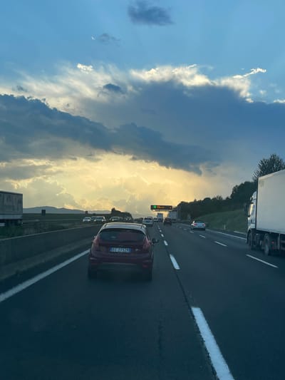The image depicts an Italian highway scene, specifically an autostrada, under a dramatic sky, likely in the vicinity of Paliano, Italy. The sky is characterized by an expanse of dark, heavy storm clouds in the upper and middle portions, contrasting with a bright golden-yellow glow emanating from the horizon where the sun is low, suggesting late afternoon or early evening. Patches of lighter blue sky are visible through the clouds, particularly in the upper left.

The perspective is from inside a vehicle, looking forward into moderate to heavy traffic. In the immediate foreground, a dark red Ford Fiesta hatchback, bearing the Italian license plate "EC 275ZM," occupies the center lane, positioned directly ahead of the viewer's vehicle. To the right, a large white semi-truck is partially visible in the rightmost lane. Further down the highway, numerous other vehicles, including sedans and hatchbacks, are visible in multiple lanes, stretching into the distance. On the far left shoulder, a white truck trailer is visible.

The highway features multiple lanes separated by dashed white lines, with a solid white line marking the left shoulder. A concrete barrier runs along the left side of the highway, beyond which lies a flat, open area with faint hills or mountains on the distant horizon. On the right side of the highway, a grassy embankment with scattered trees is visible. An overhead green digital variable message sign is suspended above the highway in the distance, displaying unreadable information. No people are discernible in the scene.