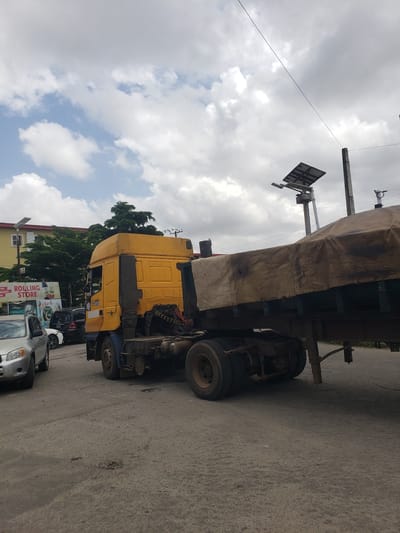 The image depicts a yellow semi-truck with a tarp-covered trailer parked on an asphalt road. The truck is positioned at an angle, with its front facing towards the right side of the frame. The trailer is long and appears to be laden. In the background, there are trees and buildings, suggesting an urban or semi-urban setting. The sky is cloudy, with patches of blue visible. The lighting suggests it is daytime, possibly overcast.
On the left side of the frame, there are some poles with what appears to be a traffic signal or a sign. There are also other vehicles parked behind the truck, including a silver car and a white car. A sign with the text "ROLLING STORE" is visible on the side of one of the buildings. The overall scene conveys a sense of everyday urban life, with the truck likely involved in transportation or delivery. The image appears to be taken from a slightly elevated perspective, looking down on the scene. The orientation of the image is rotated 90 degrees counter-clockwise from the viewer's perspective, making the truck appear to be lying on its side. The location context provided is Iganmu, Nigeria.