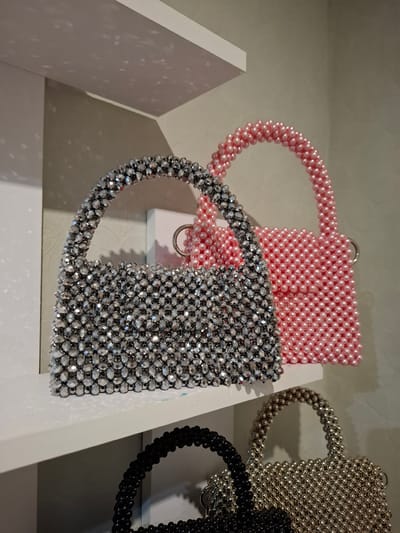 The image shows three beaded handbags displayed on white shelves. The top shelf holds two handbags, one made of silver-toned faceted beads and another of pink round beads. The handbag on the left is closer to the viewer and is fully visible, while the pink handbag on the right is partially obscured by the silver one. Below these, on a lower shelf, are two more beaded handbags, one black and one gold or bronze, both partially visible. The background is a plain, light-colored wall. The lighting casts shadows from the handbags onto the wall and shelves. The scene appears to be an indoor display, possibly in a retail setting or a home. The image was taken in Korenovsk, Russia.