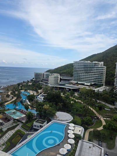 This is a high-angle, exterior shot of a resort complex situated along a rocky coastline in Jiyang District, China. The JW Marriott Hotel is prominently featured, a large, modern, multi-story building with white horizontal banding and numerous windows, backed by a lush, green, tree-covered mountainside. To the left of the hotel, a series of other hotel structures with similar architectural styles are visible. The resort grounds include several swimming pools, one with a circular pattern in its basin, and a smaller pool with what appears to be a playground structure. Palm trees and other greenery are abundant throughout the resort. The ocean stretches out to the horizon, with a few small boats visible on the water. The sky is partly cloudy with a mix of blue sky and white clouds.