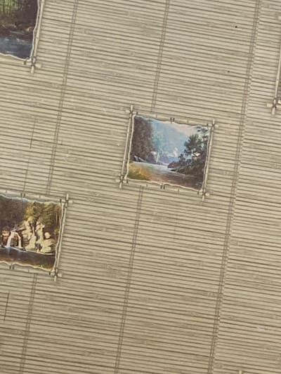 The image shows a close-up view of a bamboo mat with several small, framed landscape paintings affixed to it. The paintings depict natural scenes, including a river, trees, and a waterfall. The bamboo mat has a woven texture and appears to be a light brown or tan color. The overall impression is of decorative items in an indoor setting, possibly a home or a shop. There is no identifiable urban environment or specific location in Machara, Abkhazia discernible from the image.