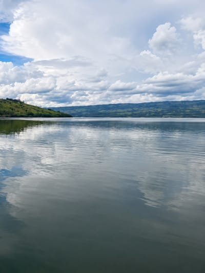 A vertical, eye-level, full shot captures a serene lake and its surroundings under a cloudy sky. The water in the foreground is calm, with gentle ripples reflecting the sky and distant landscape. The middle ground shows a rolling green hillside dotted with trees on the left, meeting a distant, hazy mountain range that stretches across the horizon. The sky above is filled with large, fluffy white clouds, with patches of blue sky visible. The scene is tranquil and natural, evoking a sense of peace. The location is identified as the city of Turpuk Sihotang, Indonesia.