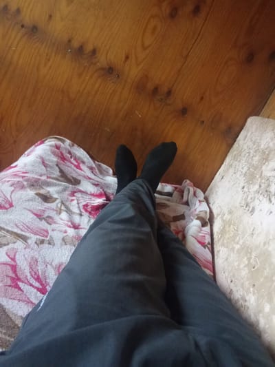 The media file shows a low-angle view of a person's legs, clad in black socks and dark grey pants, resting on a floral-patterned blanket. The blanket is predominantly white with pink and brown floral designs. To the right of the blanket, a rough, light-colored textured surface, possibly a wall or cushion, is visible. The background consists of a wooden surface with visible grain patterns and knots. The lighting suggests an indoor setting, possibly during daylight hours, with no clear indication of the time of day or weather. The scene appears to be a relaxed, personal moment, with no specific activity or event depicted beyond someone resting. There is no visible text in the image. The location context of Alatau, Kazakhstan, provides an environmental setting for this otherwise generalized domestic scene.