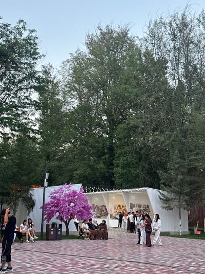 This media file depicts an outdoor event in Manas, Kyrgyzstan, likely in the late afternoon or early evening given the soft lighting. The scene is set in an open area with a patterned tiled walkway. In the background, a white tent structure is visible, adorned with string lights.
The main subjects are a group of people engaged in what appears to be a martial arts demonstration or practice. Several individuals are dressed in traditional white martial arts uniforms, some with colored belts. They are positioned in a line, suggesting a formal presentation or lesson. Other individuals are seated in the background, observing the activity.
Notable details include a vibrant artificial purple tree, adding a touch of color to the setting. Two dark-colored trash receptacles are also present. The overall atmosphere seems to be one of focused activity and polite observation. The presence of trees and the outdoor setting suggest a park or a public gathering space. No visible text is present in the image.