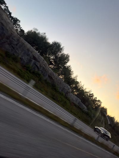 The image depicts a highway scene, likely in Rabbalshede, Sweden, during what appears to be twilight. The perspective is from a moving vehicle, indicated by motion blur on the road and surrounding landscape. A dark-colored car is visible in the right lane, also exhibiting motion blur. To the left of the highway, a steep, rocky incline covered with sparse vegetation is prominent. Dense trees line the top of the incline and extend into the background. The sky above is a gradient of pale blue to light yellow, with a few faint clouds visible. The overall impression is one of travel and the natural scenery encountered along a Swedish highway.