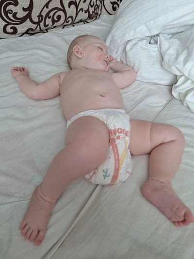 A baby is lying on its back on a white, textured surface, possibly a bed or crib. The baby is wearing a diaper. The baby's body is positioned diagonally across the frame, with its head towards the lower left and its feet towards the upper right. The baby has fair skin and appears to be very young, with chubby limbs. Redness is visible on the baby's cheeks.
The background consists of white bedding, which is wrinkled in places, suggesting a soft and comfortable environment. A portion of a patterned fabric, possibly a pillow or blanket, is visible on the far left edge of the image, featuring a brown, swirling design.
The lighting suggests it is daytime, with soft, diffused light. There are no specific weather cues or strong indications of the exact location beyond the provided context of Новобурейский, Russia. The overall impression is one of domestic tranquility and care for a young child.
The diaper the baby is wearing has visible branding. The word "HUGGIES" is clearly legible in a red font on the front of the diaper, along with some colorful graphic elements, including a yellow and red stripe pattern and a small green graphic of a stick figure.