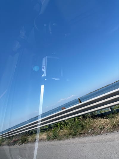 The image appears to be taken from inside a vehicle, with the windshield acting as a reflective surface. The reflection primarily shows a clear, blue sky. Outside the vehicle, visible through the windshield, is a section of road with a metal guardrail on the side. Vegetation grows along the edge of the road. Details inside the vehicle, such as parts of the dashboard and rearview mirror, are also visible in the reflection. Without more details, it is not possible to find this location in Chioggia, Italy.
