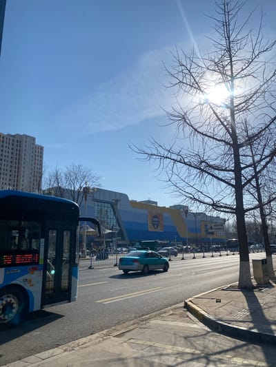 The image shows a sunny street scene in the Taihe District, China. A blue and white bus is in the foreground on the right side of the road. A light blue car is parked along the street. In the background is a large building with a yellow and blue facade, potentially a shopping center or similar commercial structure. Several other cars and what appears to be a row of shopping carts are visible next to the building. Bare trees are visible in the foreground and middle ground, casting shadows on the sidewalk. A tall building is behind the bus, opposite the main building. The sky is clear with light clouds. The street appears to be a one-way road.