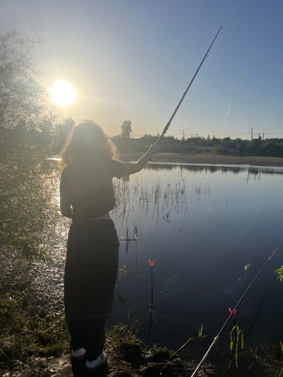 A sunlit scene features a person fishing in a body of water. The individual is silhouetted, holding a fishing rod. Another fishing rod is set up nearby. The location appears to be Krasnyi Yar, Russia, as indicated by the surrounding environment, including buildings, vegetation, and the body of water. The landscape and the structures on the opposite bank are reflected in the water's surface. The lighting indicates a sunrise or sunset.