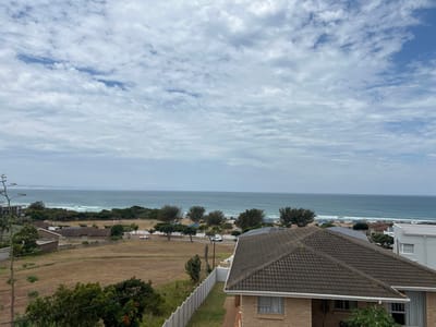 An elevated photograph captures a coastal landscape in Jeffrey's Bay, South Africa. The sky is partially cloudy, dominated by white and grey stratocumulus clouds with visible patches of blue. The Indian Ocean spans the mid-ground, displaying a light blue-grey hue with active breaking waves along the sandy shoreline. A strip of green coastal vegetation, comprising trees and bushes, runs parallel to the beach, behind which a paved road is visible. At least two vehicles, including a white sedan, are present on this road. Adjacent to the road on the left, an extensive field of dry, yellowish-brown grass extends inland. Within this field, towards the left, several low-lying structures with dark thatched roofs are discernible. In the foreground, primarily on the lower right, the dark brown tiled roof of a single-story brick residence with white gutters is prominent. The house features light brown brick walls. A light-colored fence defines a property boundary on the far right. Diverse foliage, including a tall, branched succulent-like plant on the far left, is scattered across the landscape. No human figures are visible within the frame.