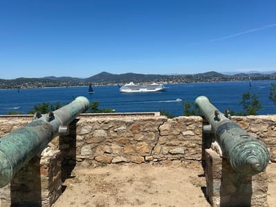 The image depicts a scenic view from a vantage point in Saint-Tropez, France, likely from the Citadel of Saint-Tropez. In the foreground, two antique bronze cannons are positioned on stone supports, overlooking a body of water. The cannons are weathered and show signs of age. Behind the cannons, a stone wall with visible cracks runs horizontally. The middle ground features a wide expanse of blue water, populated by various vessels. A large white cruise ship is prominently visible in the center, with smaller sailboats and a white speed boat also present. The background consists of a hilly coastline covered with buildings and lush greenery, under a clear, bright blue sky. The overall impression is one of historical significance combined with a beautiful coastal landscape.