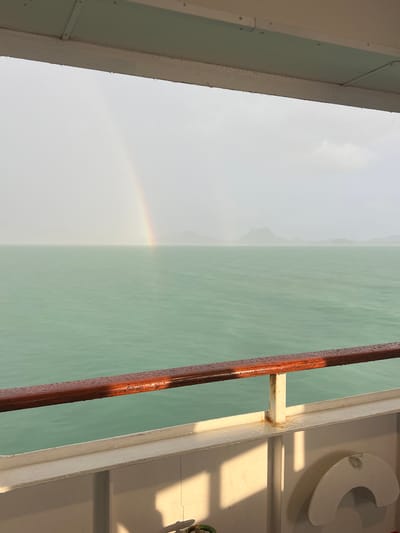 This media file depicts a view from the side of a white boat in Don Sak, Thailand. The scene is divided between a hazy sky with a faint rainbow on the left and a calm, greenish-blue body of water on the right. A dark, reddish-brown railing, appearing wet with droplets, runs vertically along the boundary between the sky and the water, suggesting it might be a handrail on the boat.
The boat's interior structure is visible on the far right, with white painted surfaces and a white C-shaped object mounted on the wall, possibly a cleat or fender holder. Sunlight casts strong shadows across the deck, indicating it is likely daytime, possibly late afternoon given the warmth of the light. The water appears placid, reflecting the overcast sky and hinting at a potentially humid or misty atmosphere, which would explain the presence of the rainbow. The overall mood is serene and tranquil, capturing a moment of natural beauty seen from the vantage point of a boat journey. There is no visible text in the image.