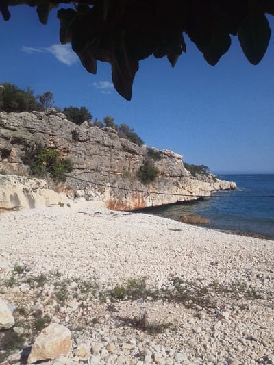 The image depicts a rocky beach in what appears to be a coastal area. The beach is covered in small, white rocks and sparse vegetation. A steep, rocky cliff rises along the back of the beach, with a mix of gray and light-colored rock formations. Some shrubs and small trees grow on top of the cliff and along its sides. The sea is visible in the background, with a clear blue sky above. There are some leaves from an overhanging tree in the top portion of the photograph. There is an unidentified cord running parallel along the cliff face.
This scene could potentially be located in or near Silifke, Turkey, given the coastal landscape and rock formations that are typical of the region. However, without more precise visual clues or identifying features, it is difficult to confirm the exact location. There are no people present in the image.