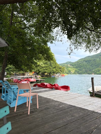 The image depicts a serene lake scene in Ćusine, Bosnia and Herzegovina, with a wooden pier in the foreground. The pier is lined with several bright red pedal boats, suggesting a recreational area. Two blue Adirondack chairs and a single pink plastic chair are placed on the pier, inviting relaxation. Beyond the pier, the calm, green water of the lake stretches out, reflecting the lush, tree-covered hills that surround it. The hills are dotted with houses, indicating a settled landscape. The sky above is overcast, contributing to the tranquil atmosphere. The overall impression is one of a peaceful, natural setting ideal for outdoor leisure activities.