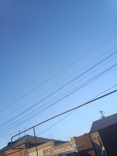 This is a low-angle, full-frame, outdoor photograph taken in Getapnya, Armenia, under a clear blue sky with no visible clouds. The image is dominated by a series of power lines that traverse diagonally across the frame, starting from the upper left and extending towards the lower right. These lines appear to be suspended between utility poles or structures that are not fully visible in the frame.

In the lower portion of the image, several man-made structures are visible. On the left, the roof of a building with a grey, corrugated surface is prominent. A rusty metal pipe runs horizontally across the middle of the image, in front of this roofline. To the right of this building, a taller structure with a darker, sloped roof is visible, possibly a shed or an extension of another building. Further to the right, a dark metal gate stands partially open, revealing a wall constructed from concrete blocks and some brickwork behind it. A faint silhouette of a transmission tower can be seen in the far upper right corner, in the sky. There are no people or animals discernible in the image. The scene suggests a residential or industrial area, characterized by utility infrastructure and various buildings.