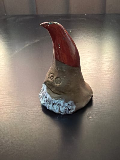 A close-up, eye-level, indoor shot shows a small, handmade ceramic gnome on a dark, textured surface. The gnome has a pointed, glossy red hat, a brown, somewhat amorphous body, a small carved face with two eyes and a curved line for a mouth, and a textured white beard with a hint of glitter. The lighting creates a slight sheen on the gnome and casts a soft shadow to its right. The background is out of focus, showing hints of a diffused, lighter colored surface above the gnome. The surface it rests on appears to be a table or countertop. The scene is devoid of any specific identifiable location in Bissendorf, Germany, as there are no external cues present.