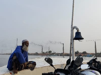 An elderly man is seated barefoot on the bow of a ferry or boat, looking right, against a hazy sky. He wears a blue long-sleeved shirt, a checkered lungi, and a white kufi cap, with a long white beard. To his right, a vertical white pole with a dark blue bell attached is visible, partially obscured by the handlebars, mirrors, and fairing of at least two parked motorcycles. The vessel appears to be traversing a river, the surface of which is visible between the boat and the distant shore.

The background across the river reveals an industrial landscape consistent with Narayanganj, Bangladesh. Multiple tall, brick-colored industrial chimneys are prominent, several emitting dark smoke plumes that rise into the pale sky, indicating active operations like brick kilns. Power transmission towers and lines are also visible in the distance, spanning across the landscape. The riverbank shows disturbed earth and scattered objects, with smaller boats or barges docked near the shore. The overall atmosphere is hazy, likely due to a combination of atmospheric conditions and industrial emissions.