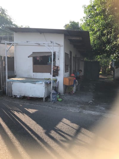 The image depicts a small, white, single-story building with a slanted, dark-colored roof. The building appears to be a shop or kiosk. In front of the building, on the left side, is a long, white chest freezer covered with a green tarp. To the right of the freezer, a small window with a brown awning is visible, displaying various items for sale. Next to the window, on the exterior wall, a patterned cloth is hanging.

Further to the right, a man in a blue shirt is seated on a stool near an open doorway. In front of the man, there is an orange crate and other items. The ground in front of the building is unpaved and appears wet in places, with some debris scattered around. A large tree with dense green foliage is visible behind and to the right of the building, casting shadows across the scene. The shadows of what appear to be the building's structure and possibly nearby trees are cast onto the asphalt road in the foreground. The overall impression is a humble, roadside establishment in a tropical environment, possibly located in Putat, Indonesia, given the vegetation and architecture.